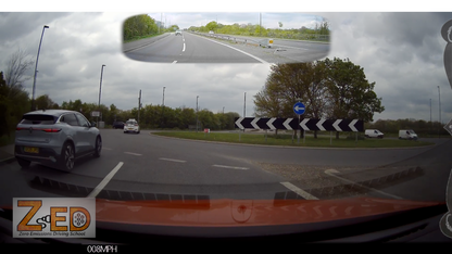 Car driving on a road with a view of the road ahead through a side mirror, showing a roundabout and trees.