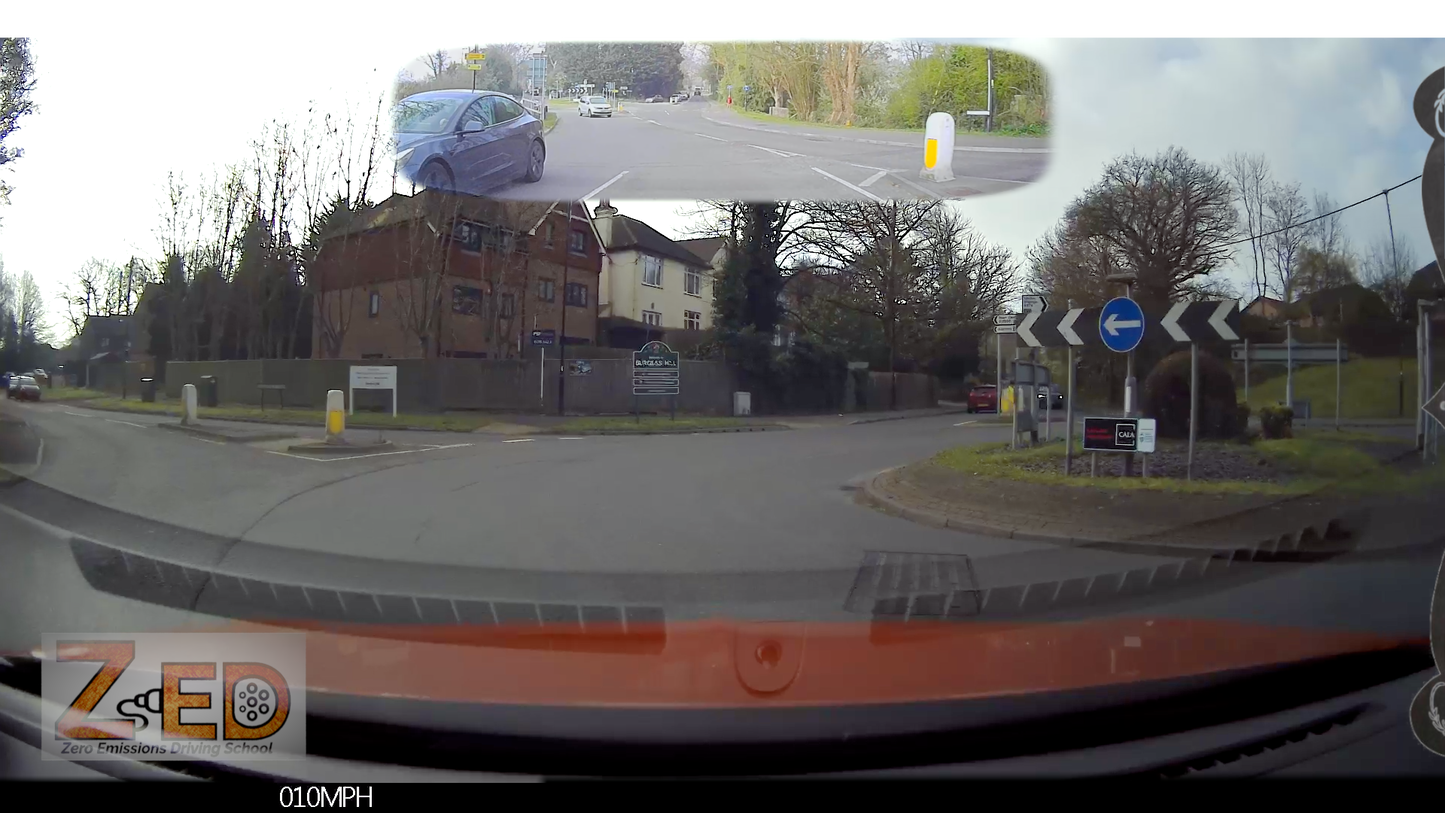 Car's front view with a ZED logo and  speedometer display, showing a road intersection with traffic lights and buildings.