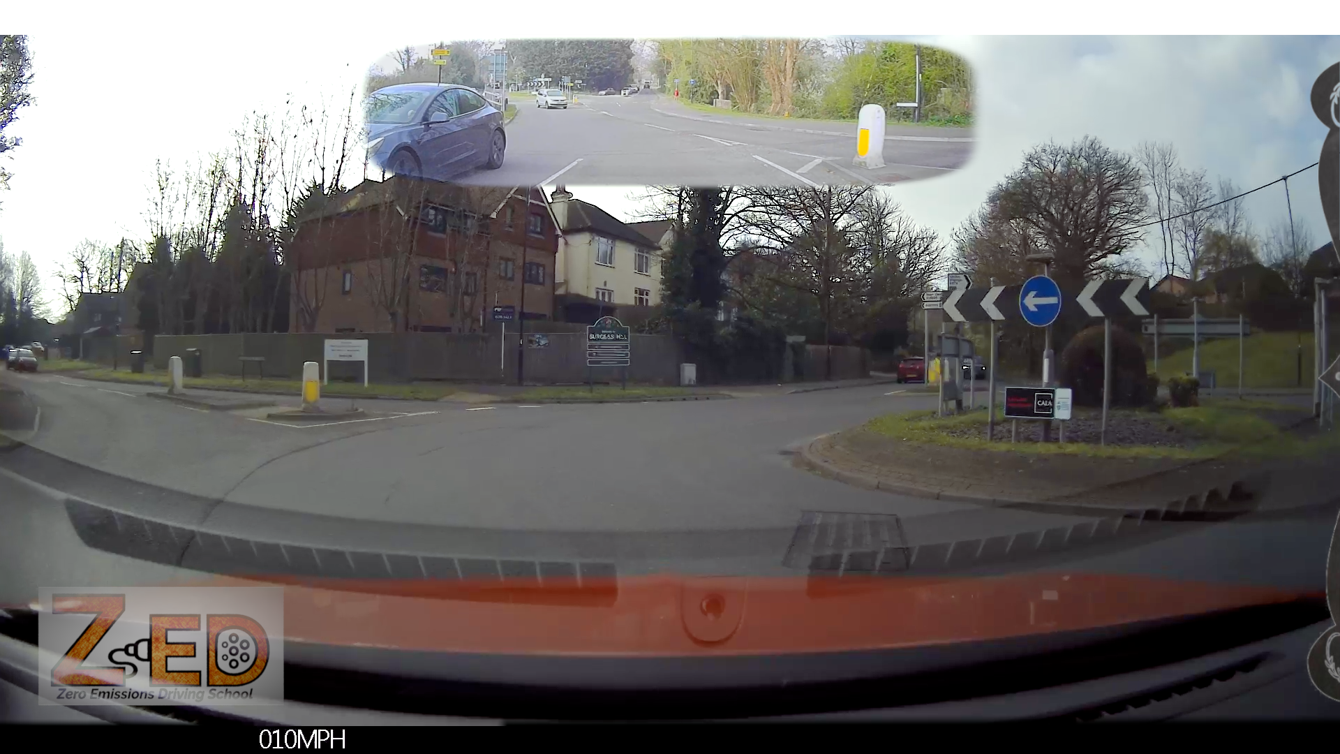 Car's front view with a ZED logo and  speedometer display, showing a road intersection with traffic lights and buildings.