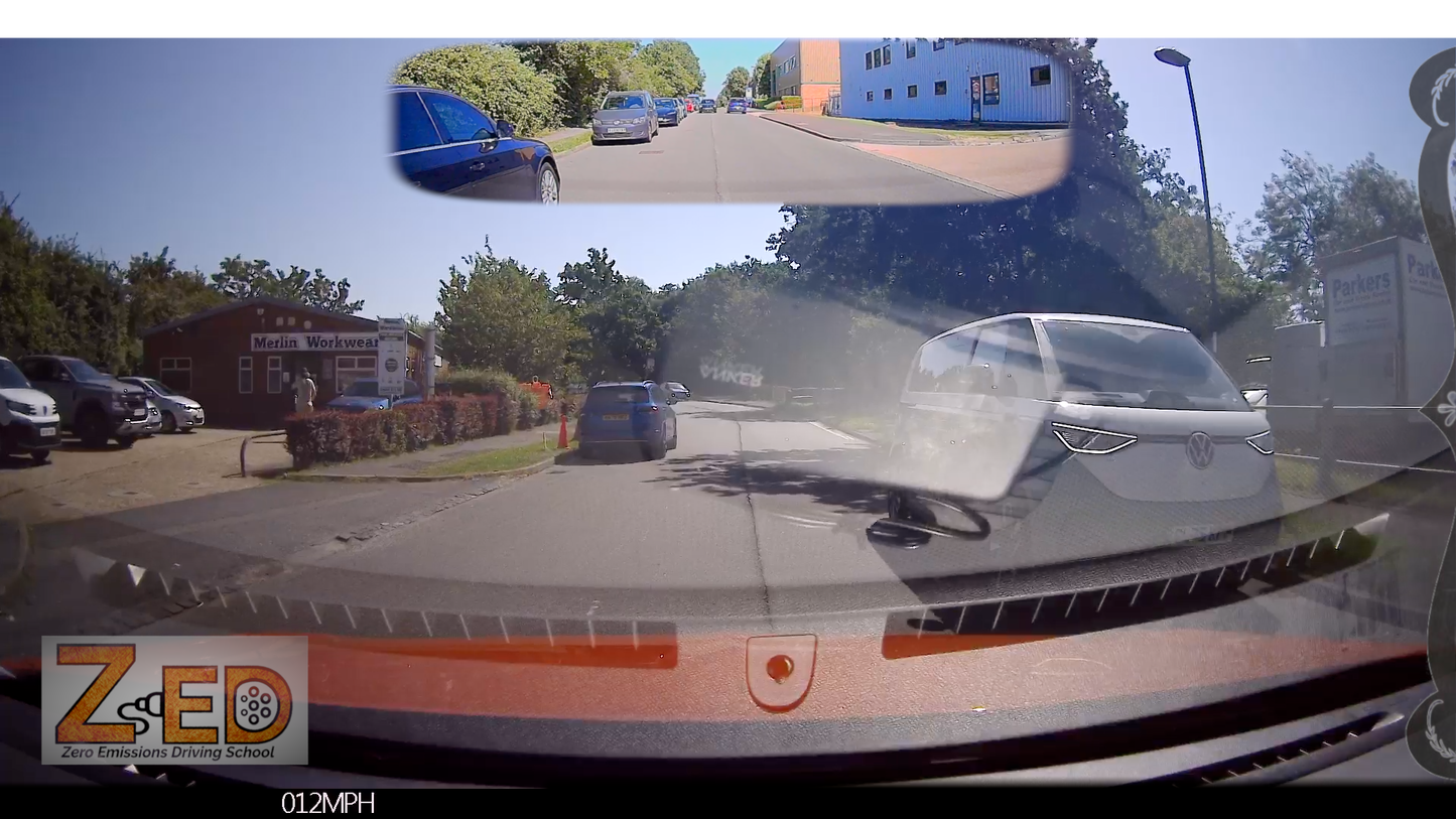 A car dashboard camera view on a sunny day, showing a road with parked and oncoming cars, trees, a blue building in the rear-view mirror, and a watermark/logo at the bottom left reading ZED.