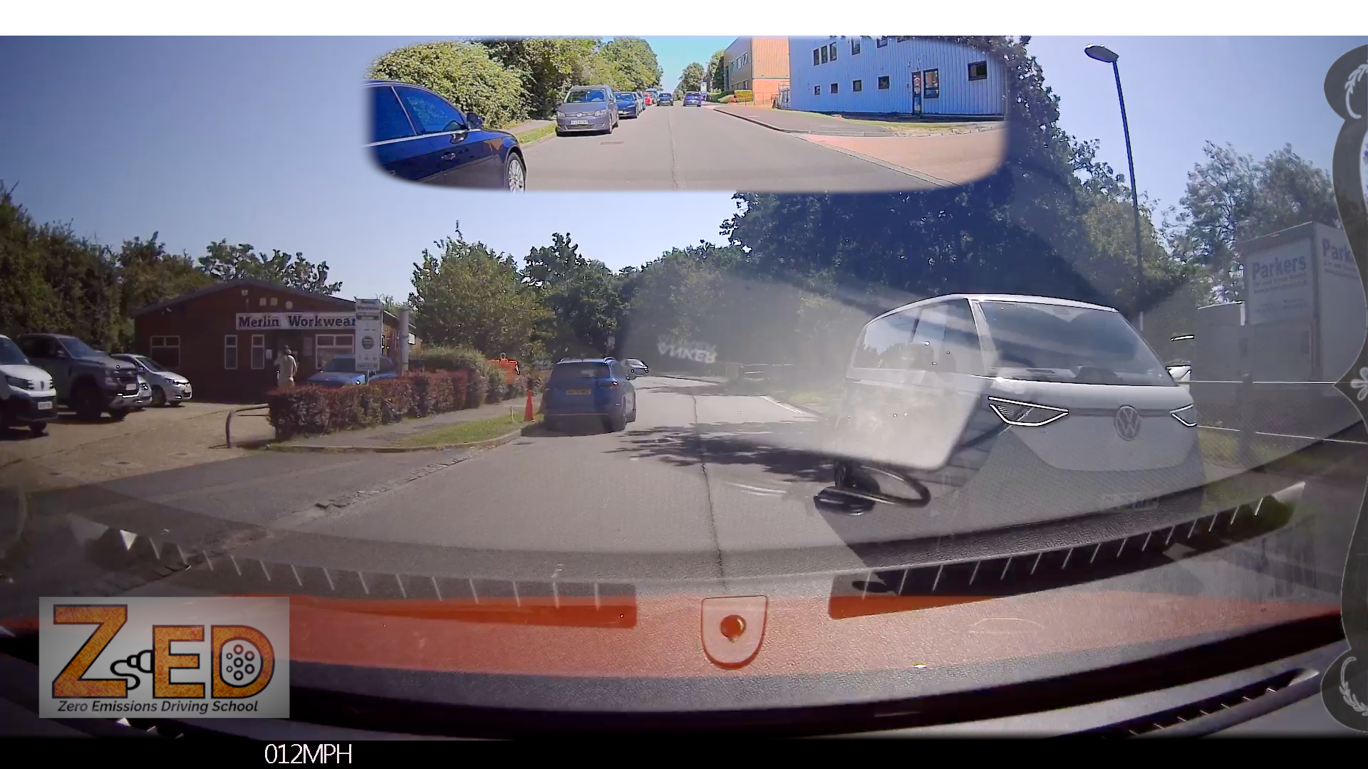 A car dashboard camera view on a sunny day, showing a road with parked and oncoming cars, trees, a blue building in the rear-view mirror, and a watermark/logo at the bottom left reading ZED.