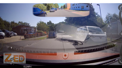 A car dashboard camera view on a sunny day, showing a road with parked and oncoming cars, trees, a blue building in the rear-view mirror, and a watermark/logo at the bottom left reading ZED.