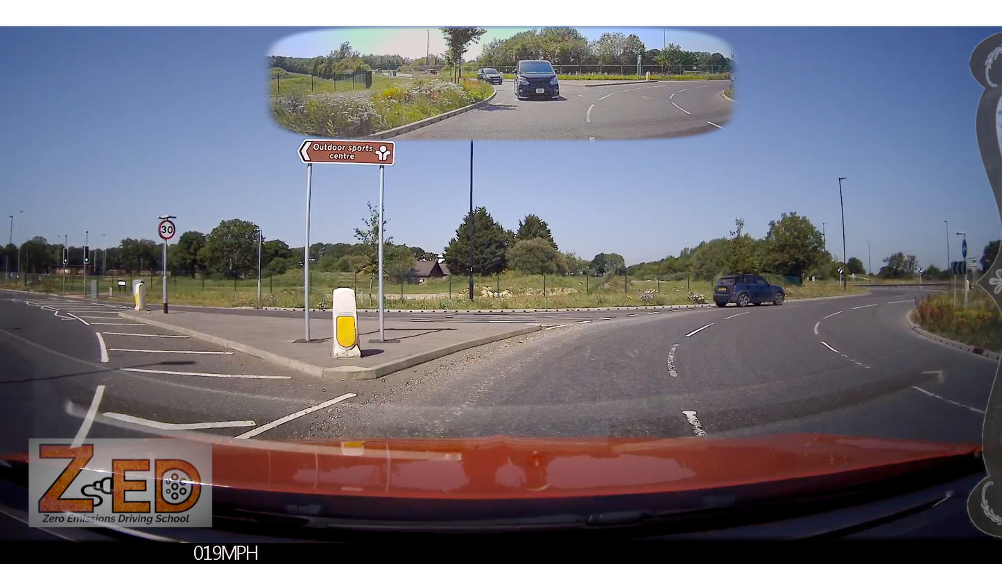 A car dashboard camera view showing a roundabout exit with a mirror inset at the top showing the rear view of a black mini van. The scene is outdoors on a sunny day with blue sky, green grass, and road signs.