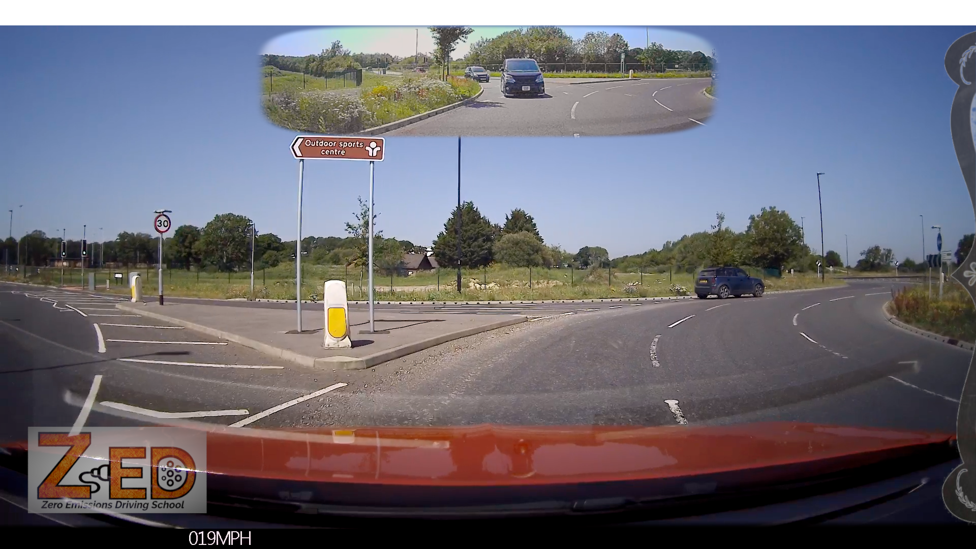 A car dashboard camera view showing a roundabout exit with a mirror inset at the top showing the rear view of a black mini van. The scene is outdoors on a sunny day with blue sky, green grass, and road signs.
