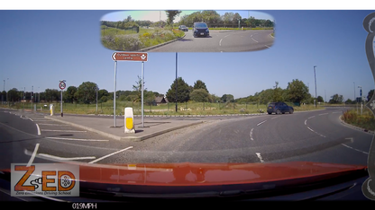 A car dashboard camera view showing a roundabout exit with a mirror inset at the top showing the rear view of a black mini van. The scene is outdoors on a sunny day with blue sky, green grass, and road signs.