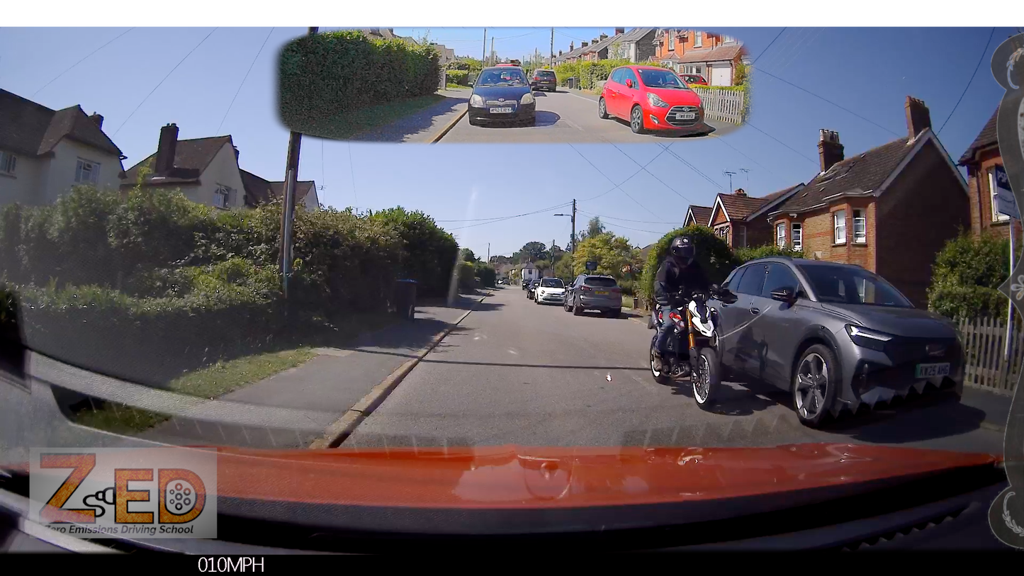 A wide-angle dashcam view from inside a car on a residential street, with a rider on a motorcycle at the front right and a red car parked on the road. A small inset at the top shows a rear-view perspective of the same street scene.