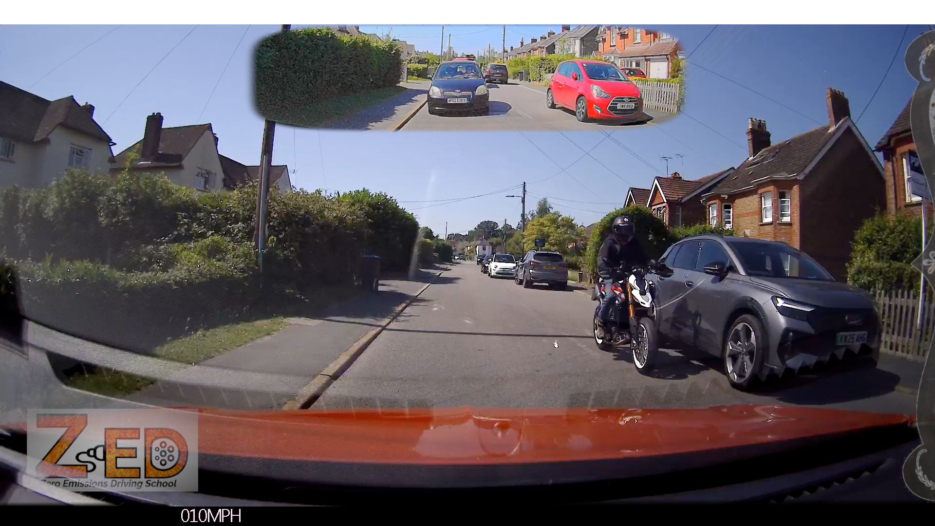 A wide-angle dashcam view from inside a car on a residential street, with a rider on a motorcycle at the front right and a red car parked on the road. A small inset at the top shows a rear-view perspective of the same street scene.