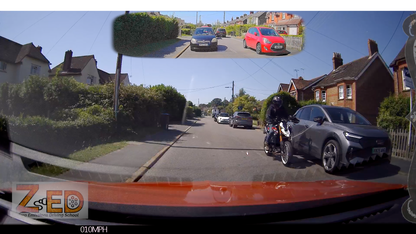 A wide-angle dashcam view from inside a car on a residential street, with a rider on a motorcycle at the front right and a red car parked on the road. A small inset at the top shows a rear-view perspective of the same street scene.