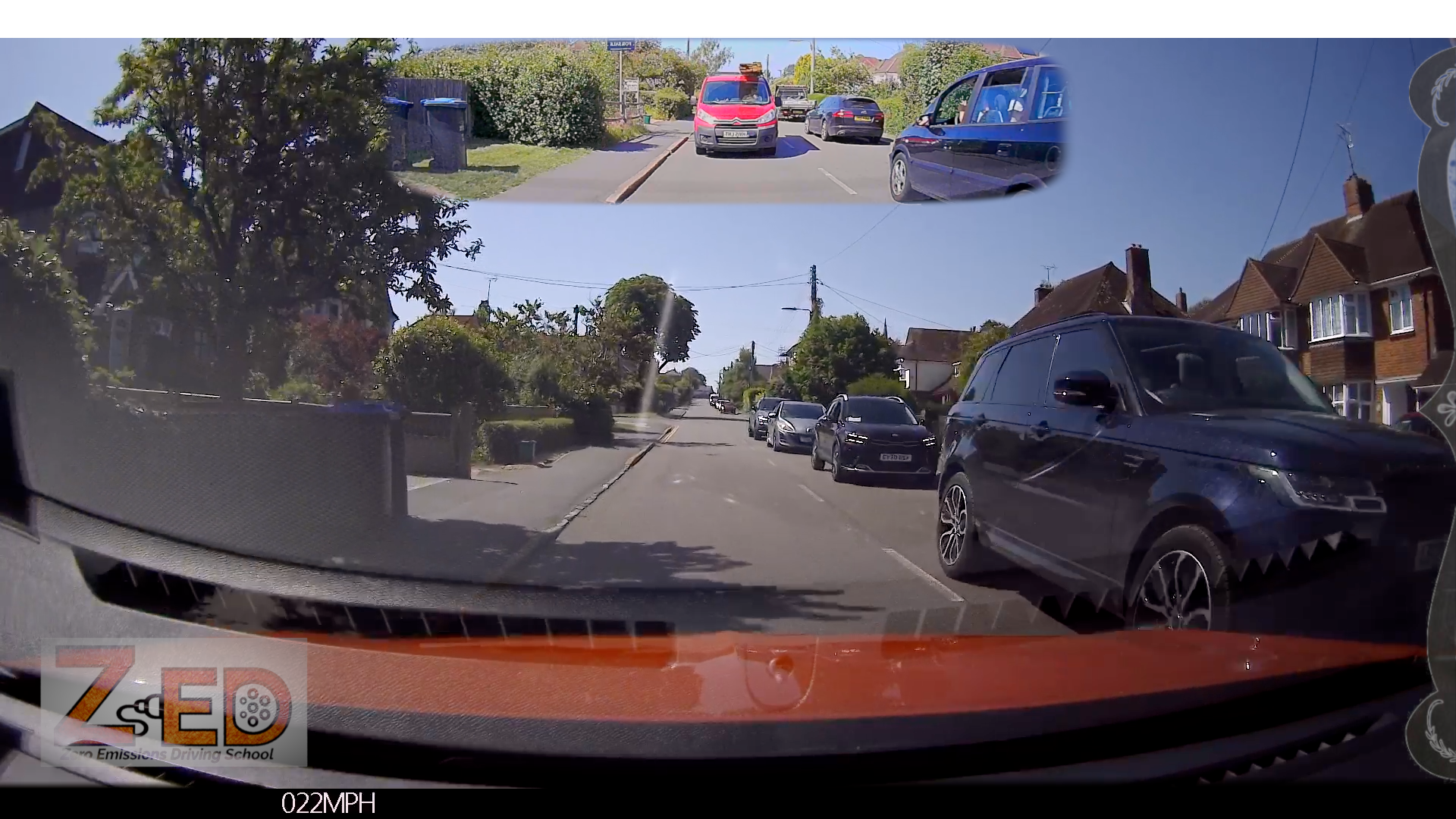 A car dashboard camera view on a residential street with houses, trees, parked cars, and a small inset at the top showing a rear-view perspective. A watermark/logo appears in the bottom left corner.