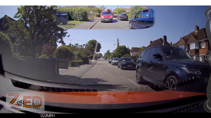 A car dashboard camera view on a residential street with houses, trees, parked cars, and a small inset at the top showing a rear-view perspective. A watermark/logo appears in the bottom left corner.