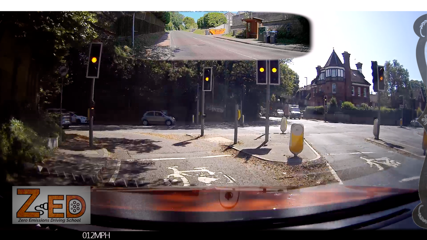 A rear-facing dashcam view from a vehicle at a road intersection, with a mirrored inset showing the view from inside the car. The scene includes traffic lights, trees, parked cars, houses, and a visible ZED School logo in the bottom left.