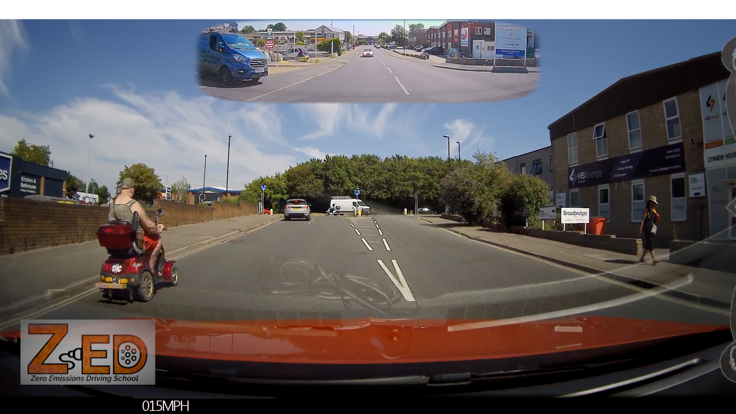 A wide-angle dashcam view from inside a vehicle on a UK road, showing the road ahead, buildings on the right, a pedestrian, and a person using a red mobility scooter on the left. A small inset at the top shows the rearview mirror perspective.
