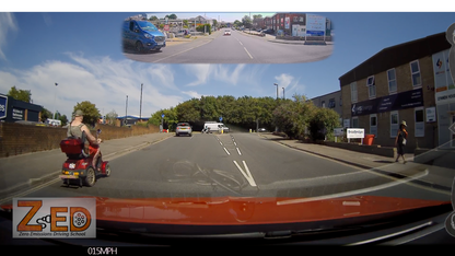 A wide-angle dashcam view from inside a vehicle on a UK road, showing the road ahead, buildings on the right, a pedestrian, and a person using a red mobility scooter on the left. A small inset at the top shows the rearview mirror perspective.