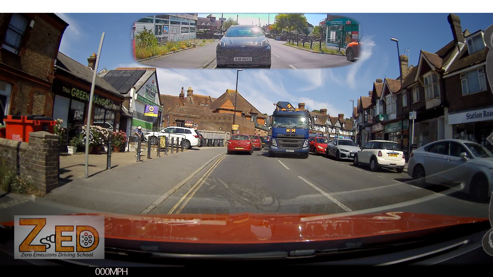 A wide-angle dashcam view from inside a vehicle on a busy street, with houses, parked cars, a blue truck, and a small inset reflection at the top showing the car's rear view. A large 'ZED' logo appears in the bottom-left corner.