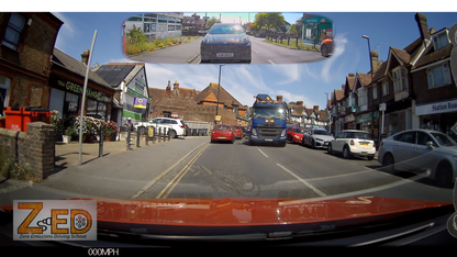 A wide-angle dashcam view from inside a vehicle on a busy street, with houses, parked cars, a blue truck, and a small inset reflection at the top showing the car's rear view. A large 'ZED' logo appears in the bottom-left corner.