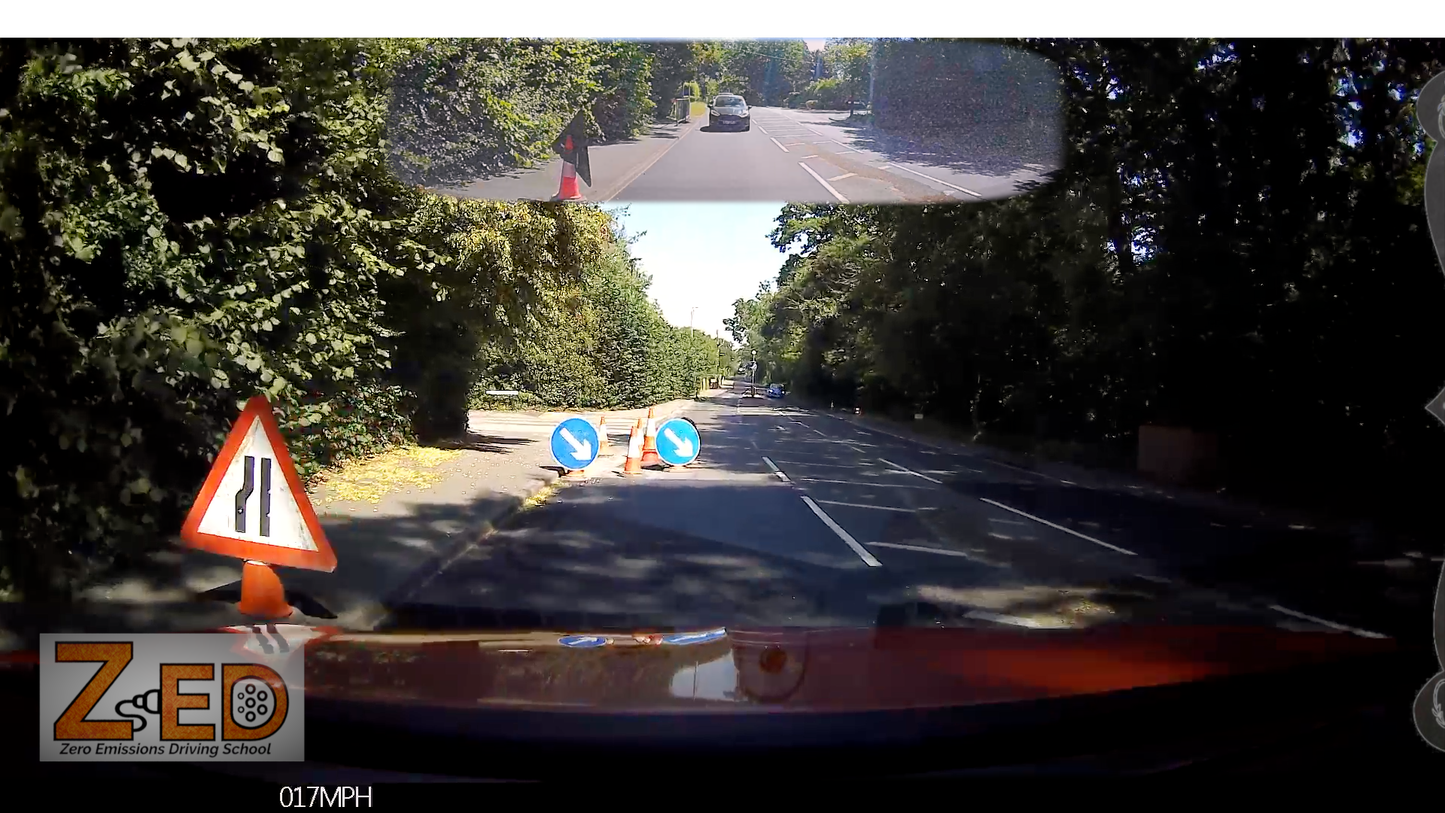 A car dashboard camera view on a tree-lined road with road work ahead, showing a speed limit sign, directional arrows, a warning triangle, and a rearview mirror reflection of the road behind.