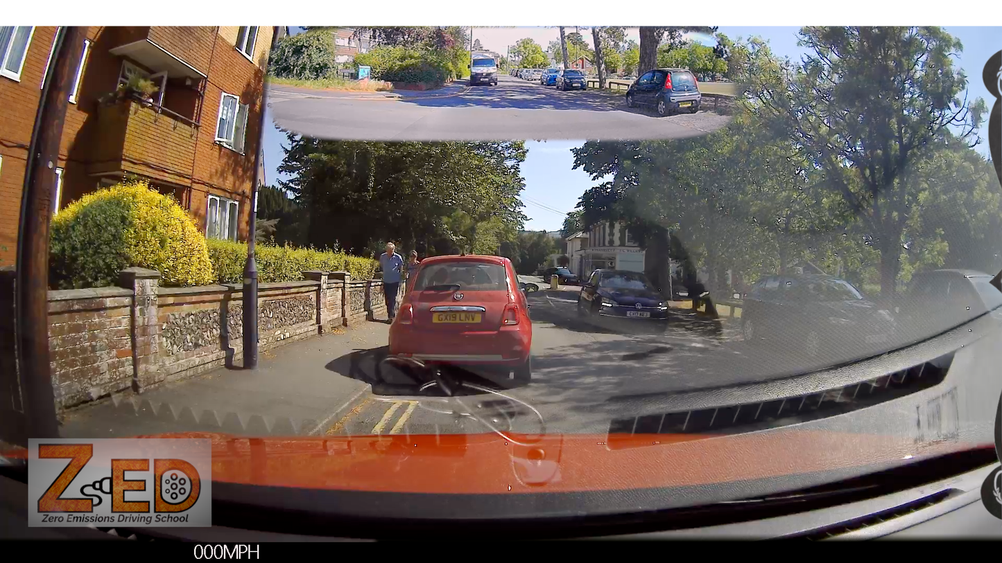 A car dashcam view on a residential street, showing a red car in front, parked cars on both sides, trees, and a building on the left. The image has a reflective glare on the windshield and a logo watermark in the bottom-left corner.
