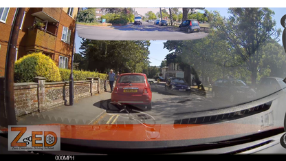 A car dashcam view on a residential street, showing a red car in front, parked cars on both sides, trees, and a building on the left. The image has a reflective glare on the windshield and a logo watermark in the bottom-left corner.